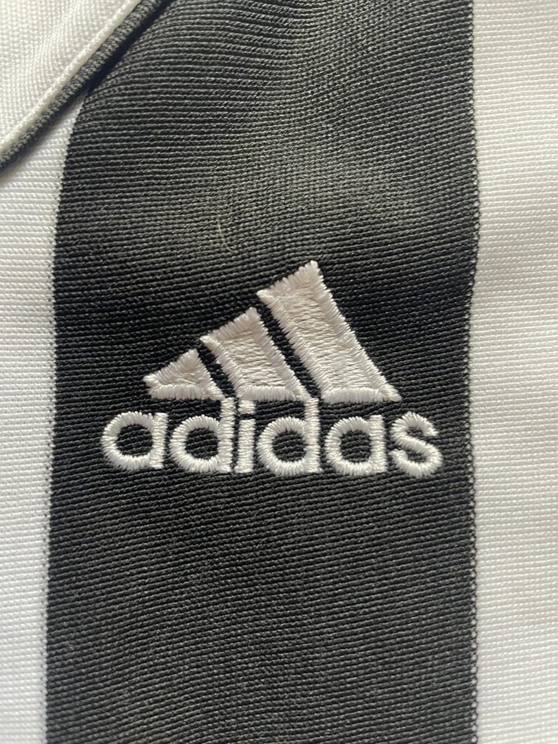May include: Close-up of a black and white striped fabric with the Adidas logo embroidered in white. The Adidas logo is a white trefoil with the word "adidas" stitched below it.