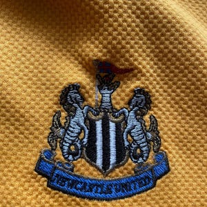 May include: A yellow soccer jersey with the Newcastle United crest embroidered on the front. The crest features a black and white shield with two black and white seahorses on either side.