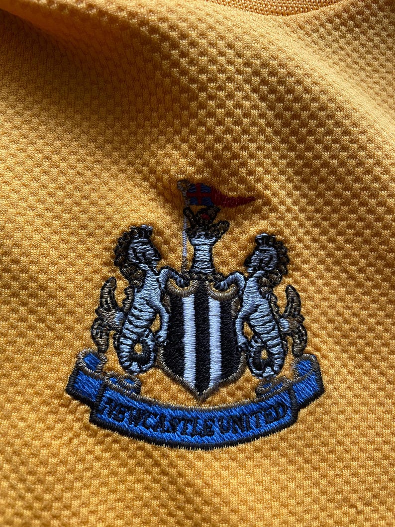 May include: A yellow soccer jersey with the Newcastle United crest embroidered on the front. The crest features a black and white shield with two black and white seahorses on either side.