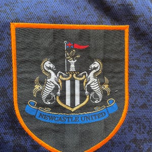 May include: Close-up of a Newcastle United crest patch. The patch features a shield shape with an orange border, a black background, and the club's emblem. The emblem includes two seahorses, a crest, and the words "Newcastle United".