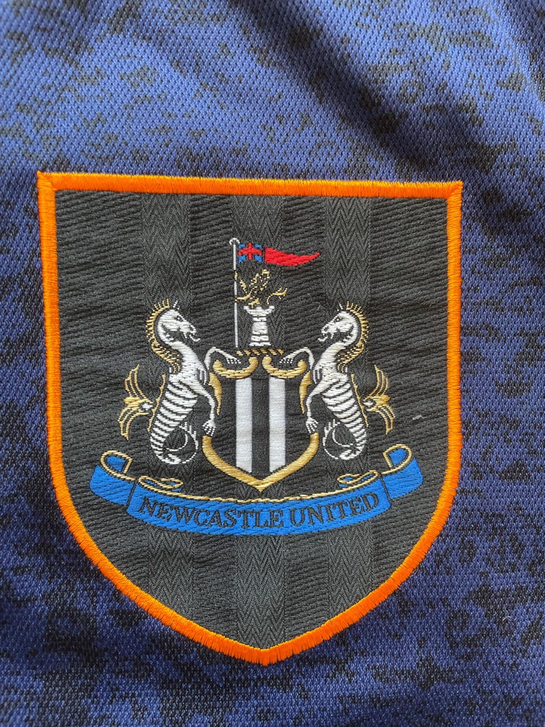 May include: Close-up of a Newcastle United crest patch. The patch features a shield shape with an orange border, a black background, and the club's emblem. The emblem includes two seahorses, a crest, and the words "Newcastle United".