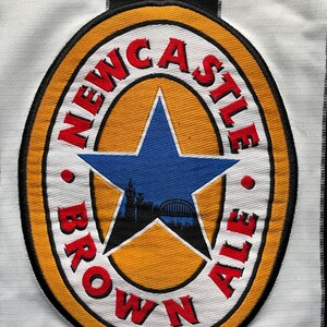 May include: A white and black striped soccer jersey with a Newcastle Brown Ale logo patch. The logo is a yellow circle with a red star and the words "Newcastle Brown Ale" in red.
