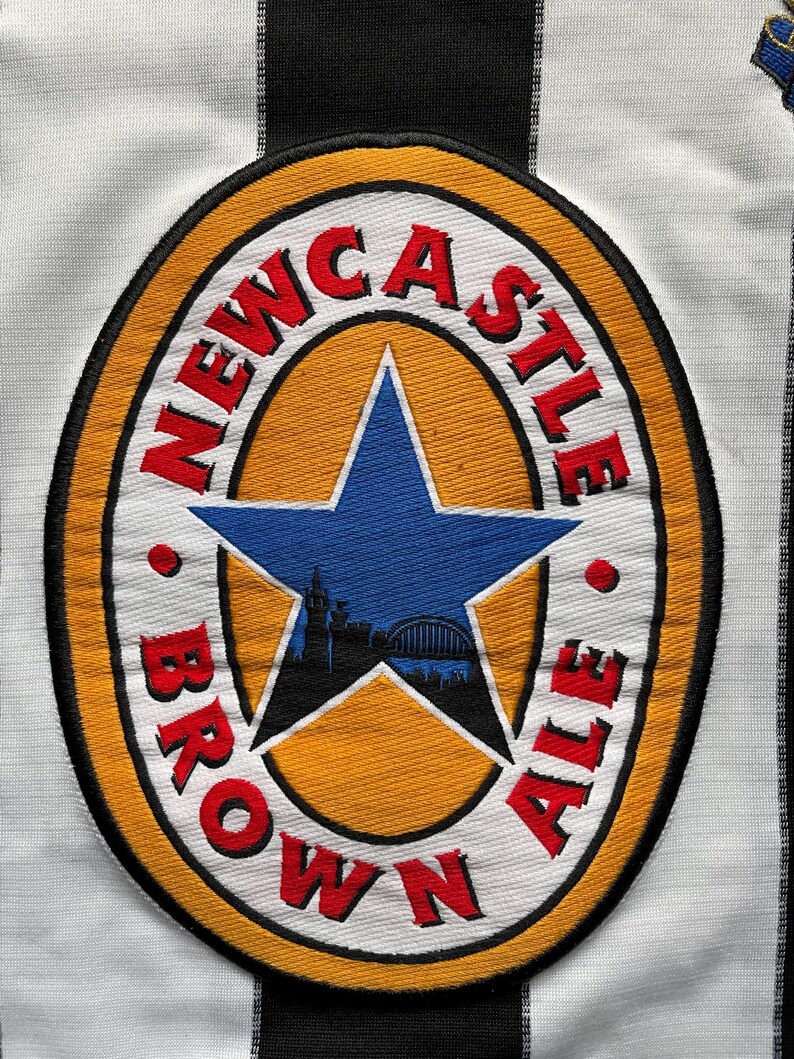 May include: A white and black striped soccer jersey with a Newcastle Brown Ale logo patch. The logo is a yellow circle with a red star and the words "Newcastle Brown Ale" in red.