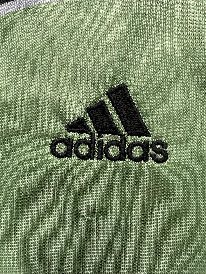 May include: A close-up of a green Adidas logo with black stitching. The logo is embroidered on a green fabric.