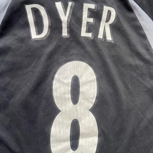 以下が含まれることがあります： 黒のサッカーユニフォーム。背中に「DYER」と「8」の番号が書かれている。ユニフォームにはFAプレミアリーグのロゴがある。