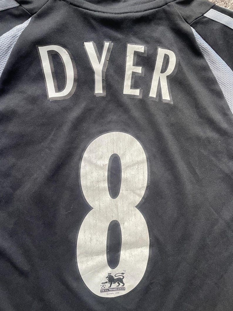 以下が含まれることがあります： 黒のサッカーユニフォーム。背中に「DYER」と「8」の番号が書かれている。ユニフォームにはFAプレミアリーグのロゴがある。