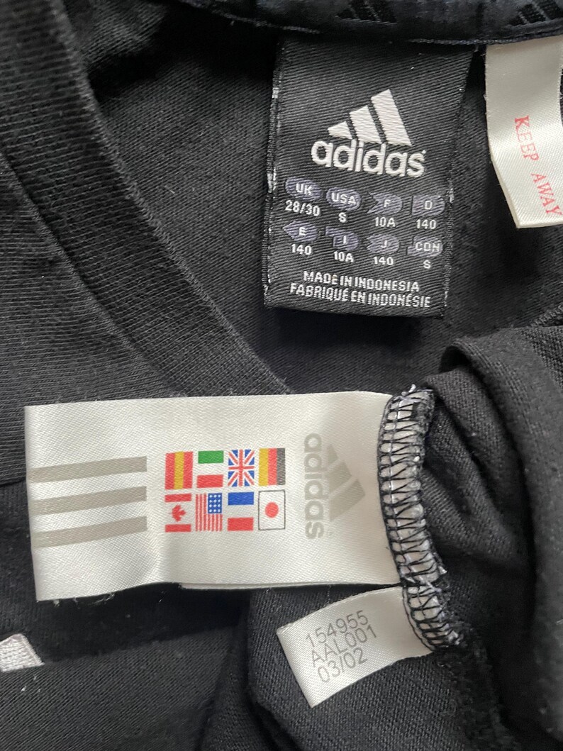 May include: Black Adidas t-shirt with a white Adidas logo and size tag. The tag shows sizes in UK, USA, EU, and J, and the text "Made in Indonesia, Fabriqu&eacute; en Indon&eacute;sie".