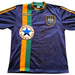 May include: A navy blue soccer jersey with green and orange accents. The jersey features the Adidas logo, a Newcastle Brown Ale patch, and a team crest. The sleeves and collar are green, with orange stripes on the shoulders.