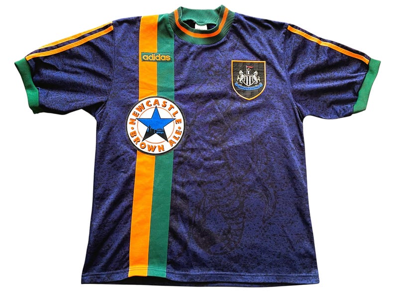 May include: A navy blue soccer jersey with green and orange accents. The jersey features the Adidas logo, a Newcastle Brown Ale patch, and a team crest. The sleeves and collar are green, with orange stripes on the shoulders.
