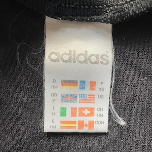 May include: Close-up of a black garment tag with the Adidas logo in white. The tag includes size and country codes, such as US, GB, and CDN, along with corresponding flags. The garment appears to be a small size.