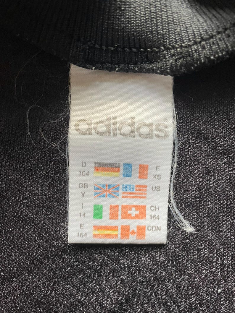 May include: Close-up of a black garment tag with the Adidas logo in white. The tag includes size and country codes, such as US, GB, and CDN, along with corresponding flags. The garment appears to be a small size.