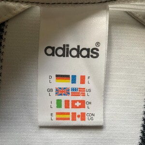 May include: Close-up of a white Adidas tag with the brand's logo in black. The tag includes flags representing various countries, including Germany, France, the United Kingdom, and the United States.