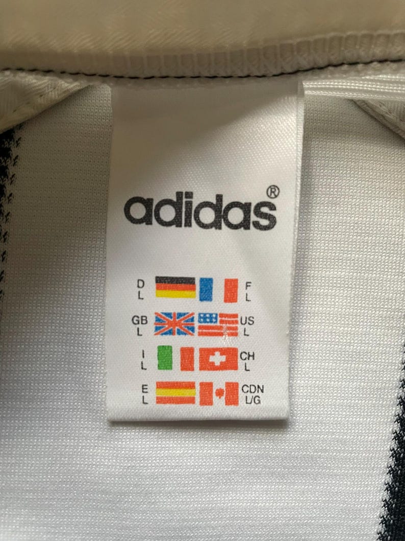 May include: Close-up of a white Adidas tag with the brand's logo in black. The tag includes flags representing various countries, including Germany, France, the United Kingdom, and the United States.