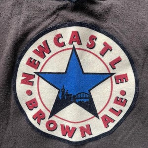 Puede incluir: Primer plano de una tela gris oscuro con dos parches bordados. El parche m&aacute;s grande presenta una estrella azul, las palabras "NEWCASTLE BROWN ALE" en rojo y una silueta de un puente y edificios. El parche m&aacute;s peque&ntilde;o tiene las palabras "NEWCASTLE UNITED".