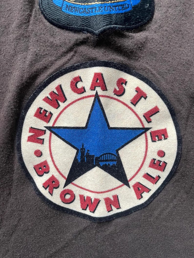 Puede incluir: Primer plano de una tela gris oscuro con dos parches bordados. El parche m&aacute;s grande presenta una estrella azul, las palabras "NEWCASTLE BROWN ALE" en rojo y una silueta de un puente y edificios. El parche m&aacute;s peque&ntilde;o tiene las palabras "NEWCASTLE UNITED".