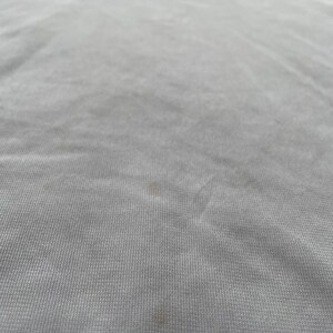Pode incluir: Uma camiseta cinza claro com a palavra "ROCK" impressa em letras douradas na parte superior. O tecido tem uma textura sutil e algumas pequenas manchas marrons s&atilde;o vis&iacute;veis.