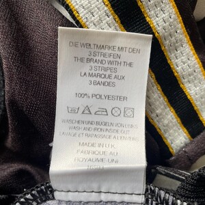 May include: Close-up of a white tag with text in multiple languages, including German, English, and French. The tag reads "THE BRAND WITH THE 3 STRIPES" and "100% POLYESTER." The tag is attached to a black and white patterned fabric with yellow stripes.