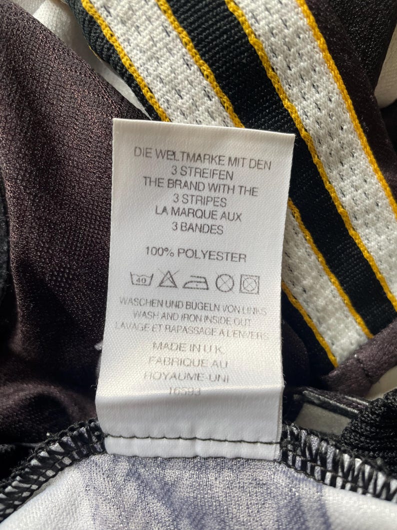 May include: Close-up of a white tag with text in multiple languages, including German, English, and French. The tag reads "THE BRAND WITH THE 3 STRIPES" and "100% POLYESTER." The tag is attached to a black and white patterned fabric with yellow stripes.