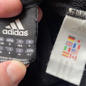 May include: Black adidas sweatpants with a black tag that says "adidas" and has size information in inches and centimetres. A white tag with the adidas logo and flags of different countries is attached to the sweatpants.