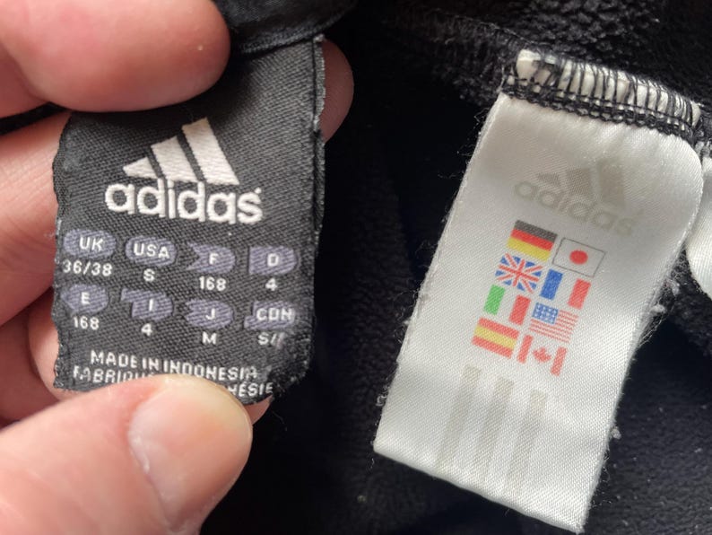 May include: Black adidas sweatpants with a black tag that says "adidas" and has size information in inches and centimetres. A white tag with the adidas logo and flags of different countries is attached to the sweatpants.