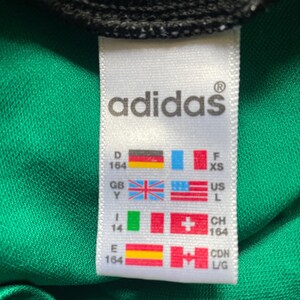 K&ouml;nnte beinhalten: Nahaufnahme eines wei&szlig;en Adidas-Etiketts mit Markennamen und Gr&ouml;&szlig;enangaben. Das Etikett befindet sich auf einem gr&uuml;nen Stoff mit schwarzem Besatz oben. Das Etikett enth&auml;lt Flaggen verschiedener L&auml;nder.
