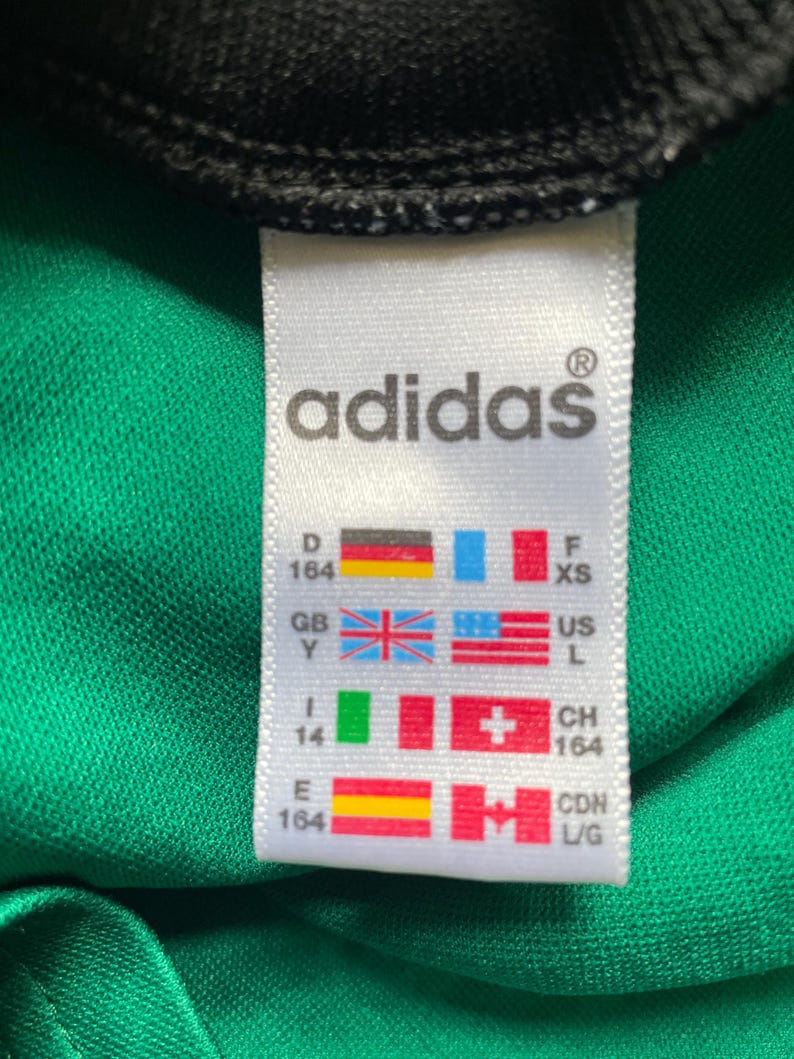 K&ouml;nnte beinhalten: Nahaufnahme eines wei&szlig;en Adidas-Etiketts mit Markennamen und Gr&ouml;&szlig;enangaben. Das Etikett befindet sich auf einem gr&uuml;nen Stoff mit schwarzem Besatz oben. Das Etikett enth&auml;lt Flaggen verschiedener L&auml;nder.