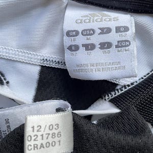 Pode incluir: Camisola de futebol branca e preta com riscas com um log&oacute;tipo Adidas branco e uma etiqueta de tamanho. A etiqueta mostra os tamanhos em Reino Unido, EUA, F e D. A etiqueta tamb&eacute;m diz "Made in Bulgaria, Fabrique en Bulgarie". Existe uma etiqueta branca com o texto "12/03 021786 CRA001".