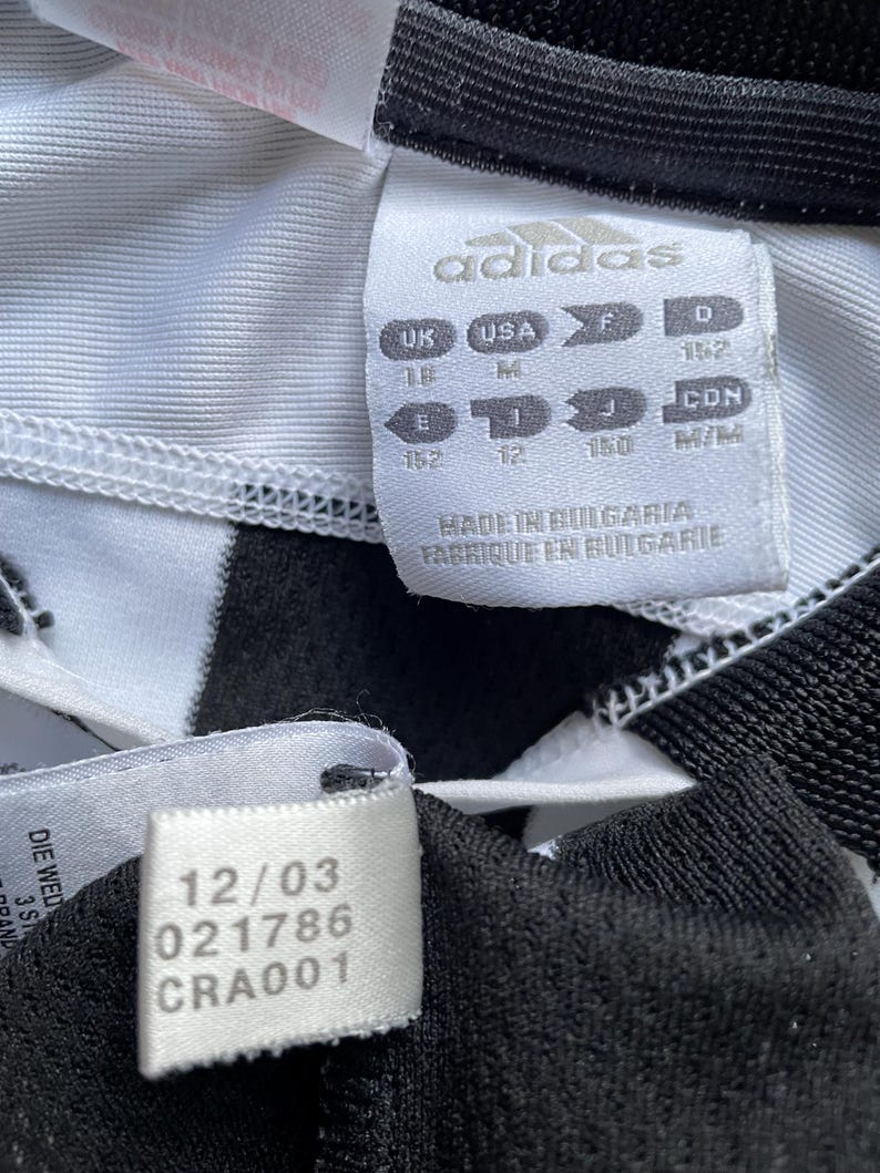 Pode incluir: Camisola de futebol branca e preta com riscas com um log&oacute;tipo Adidas branco e uma etiqueta de tamanho. A etiqueta mostra os tamanhos em Reino Unido, EUA, F e D. A etiqueta tamb&eacute;m diz "Made in Bulgaria, Fabrique en Bulgarie". Existe uma etiqueta branca com o texto "12/03 021786 CRA001".