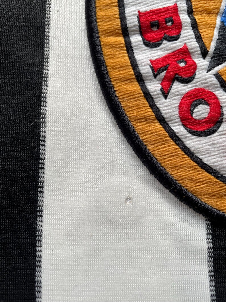 Puede incluir: Camiseta de f&uacute;tbol a rayas blancas y negras con un parche bordado amarillo, negro y rojo que dice "BRO" en rojo.