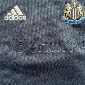 Puede incluir: Una camiseta de f&uacute;tbol azul marino con detalles blancos. La camiseta tiene el logotipo de Adidas en el pecho izquierdo y el escudo del Newcastle United en el pecho derecho. El texto "home" est&aacute; bordado en blanco en la parte delantera de la camiseta.