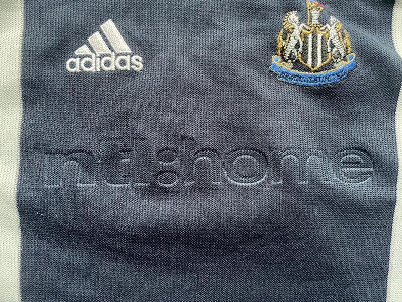 Puede incluir: Una camiseta de f&uacute;tbol azul marino con detalles blancos. La camiseta tiene el logotipo de Adidas en el pecho izquierdo y el escudo del Newcastle United en el pecho derecho. El texto "home" est&aacute; bordado en blanco en la parte delantera de la camiseta.