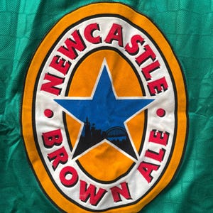May include: Close-up of a green fabric with a circular logo. The logo features the words "NEWCASTLE BROWN ALE" in red, surrounding a blue star with a city silhouette. The logo is set against an orange background.