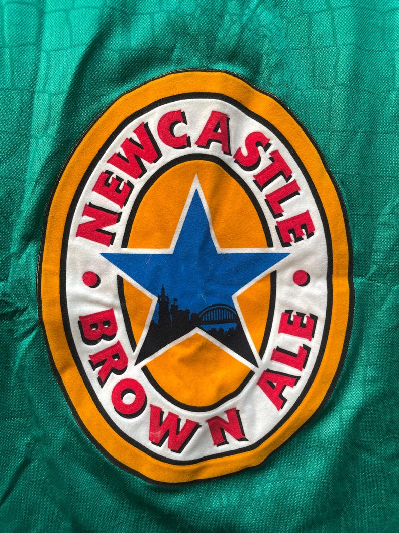 May include: Close-up of a green fabric with a circular logo. The logo features the words "NEWCASTLE BROWN ALE" in red, surrounding a blue star with a city silhouette. The logo is set against an orange background.