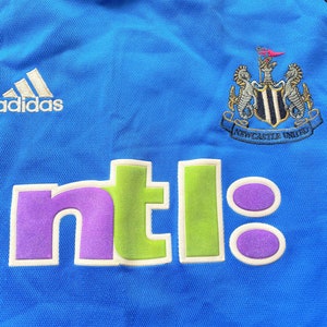 May include: A blue soccer jersey with the Newcastle United logo and the word "ntl" in green, purple, and white. The Adidas logo is on the left side of the jersey.