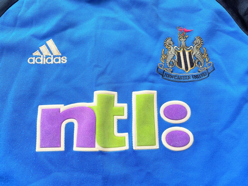 May include: A blue soccer jersey with the Newcastle United logo and the word "ntl" in green, purple, and white. The Adidas logo is on the left side of the jersey.