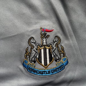 May include: A gray embroidered patch with a black and white striped shield, two gold seahorses, and a blue ribbon with the text "Newcastle United".