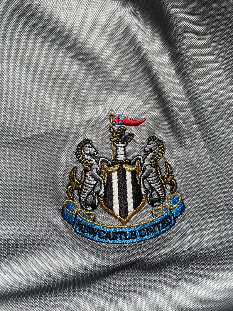 May include: A gray embroidered patch with a black and white striped shield, two gold seahorses, and a blue ribbon with the text "Newcastle United".