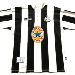 May include: A black and white striped soccer jersey with the Adidas logo. The jersey features a Newcastle Brown Ale logo and a team crest. The shirt has a collared neckline and button closure.