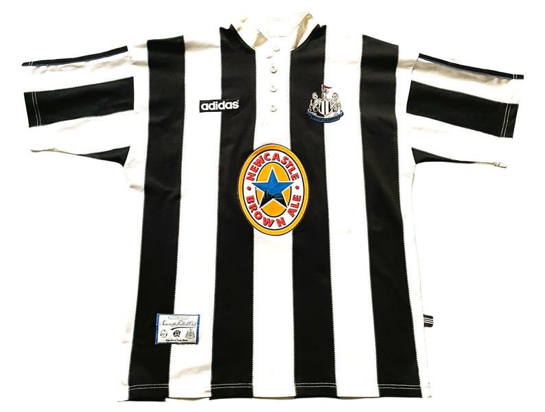 May include: A black and white striped soccer jersey with the Adidas logo. The jersey features a Newcastle Brown Ale logo and a team crest. The shirt has a collared neckline and button closure.