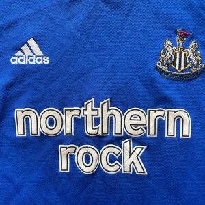May include: A blue soccer jersey with white Adidas logo, a Newcastle United crest, and the words "northern rock" in white.