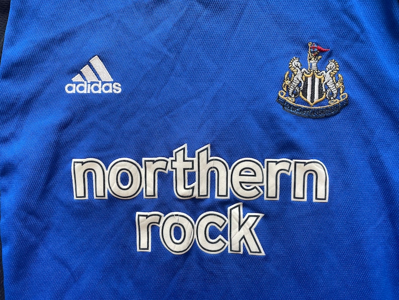 May include: A blue soccer jersey with white Adidas logo, a Newcastle United crest, and the words "northern rock" in white.