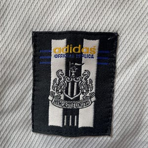 Pu&ograve; includere: Una toppa bianca e nera con il logo adidas e il testo "Official Replica" in giallo. La toppa presenta anche lo stemma del Newcastle United.