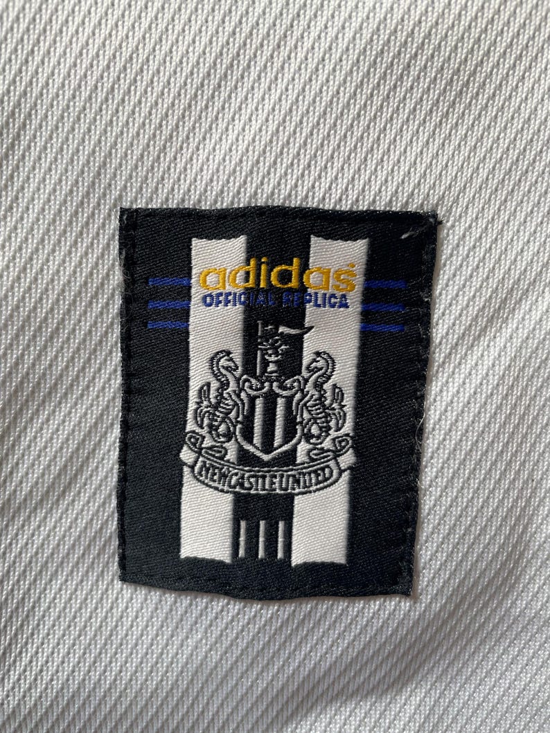 Pu&ograve; includere: Una toppa bianca e nera con il logo adidas e il testo "Official Replica" in giallo. La toppa presenta anche lo stemma del Newcastle United.