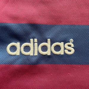 May include: Close-up of a pink and blue striped fabric with the embroidered word "adidas" in white. The fabric has a textured appearance, with the stripes running horizontally across the image. The Adidas logo is centered.