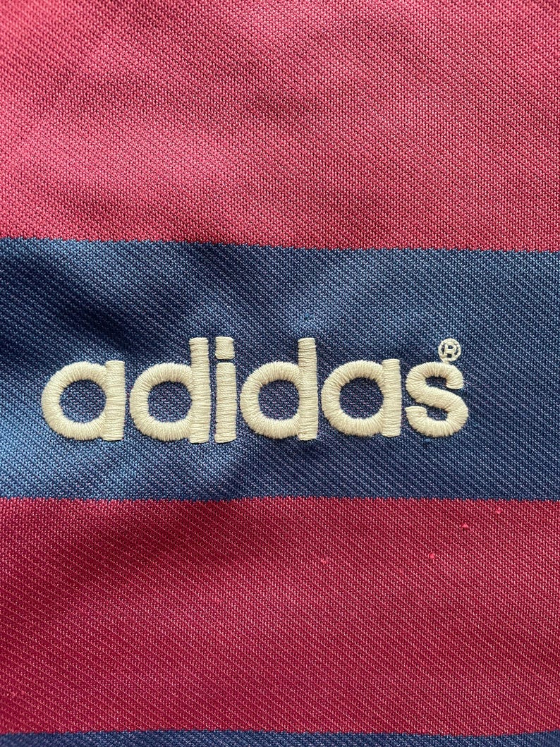 May include: Close-up of a pink and blue striped fabric with the embroidered word "adidas" in white. The fabric has a textured appearance, with the stripes running horizontally across the image. The Adidas logo is centered.