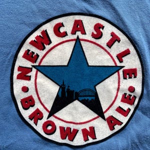 May include: A blue t-shirt with a white circle logo featuring a red and black star with a city skyline inside. The text around the star reads "NEWCASTLE BROWN ALE."