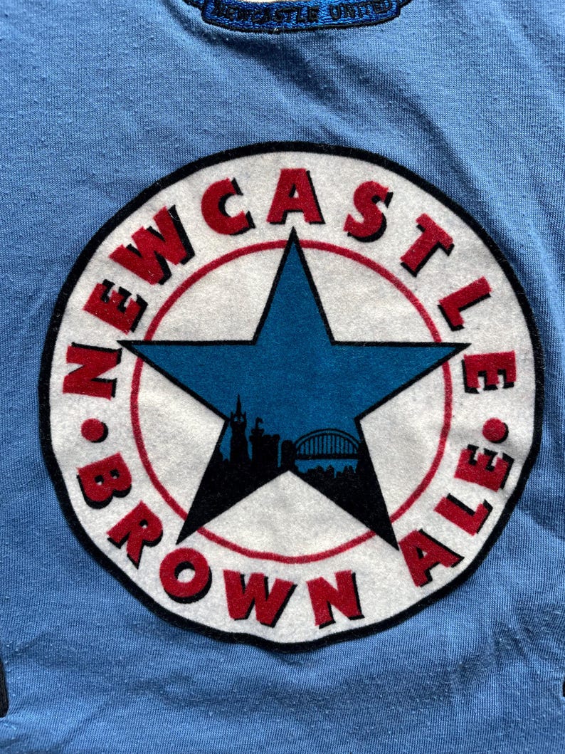 May include: A blue t-shirt with a white circle logo featuring a red and black star with a city skyline inside. The text around the star reads "NEWCASTLE BROWN ALE."