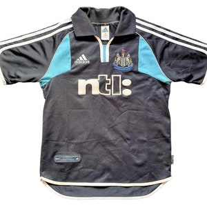 Op de afbeelding: Een zwart voetbaltenue met witte en blauwe accenten. Het tenue heeft het Adidas-logo, het Newcastle United-wapen en het NTL-logo. Het tenue is gemaakt van een lichtgewicht, ademende stof.