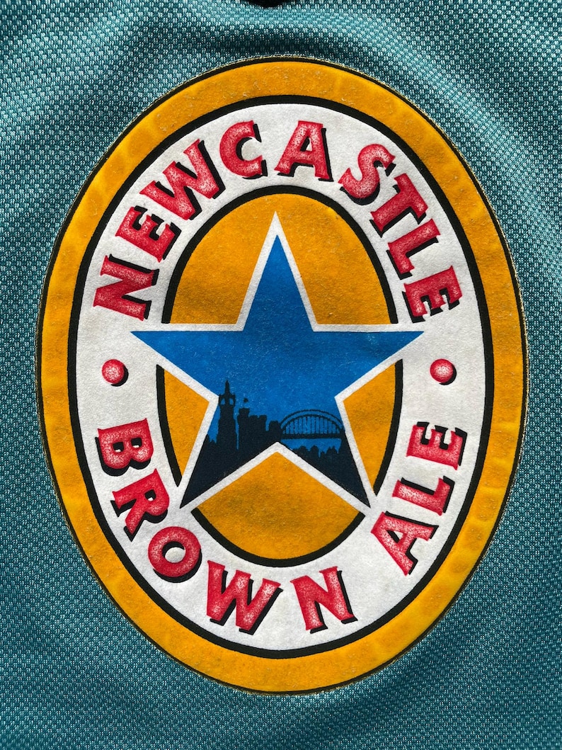 Pu&ograve; includere: Una maglia color teal con il logo Newcastle Brown Ale. Il logo ovale presenta uno sfondo giallo, una stella blu e la scritta "NEWCASTLE BROWN ALE" in rosso. La maglia ha un motivo strutturato.