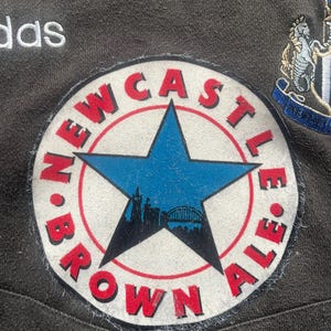 May include: A white and red circular patch with the text "NEWCASTLE BROWN ALE" around a blue five-pointed star with a cityscape inside. The star is on a white background.